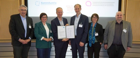 Sechs Personen stehen vor einer Präsentationswand, zwei davon halten ein Dokument im Rahmen einer offiziellen Unterzeichnung in die Kamera.