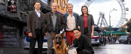 Vier Personen und ein Schäferhund posieren bei Dreharbeiten im Wiener Prater vor einem Polizeiauto und dem Riesenrad; im Vordergrund hält ein Crewmitglied eine Filmklappe.