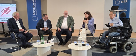 Expert:innen diskutieren auf einem Podium über Barrierefreiheit in audiovisuellen Medien. Ein Teilnehmer nimmt im Elektrorollstuhl an der Diskussion teil.