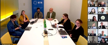 Mehrere Vertreter:innen der RTR-GmbH sitzen an einem Besprechungstisch und begrüßen die Teilnehmenden des Medienkompetenz-Roundtables. Die Veranstaltung findet hybrid statt, weitere Teilnehmende sind rechts in der Videokonferenz eingeblendet.
