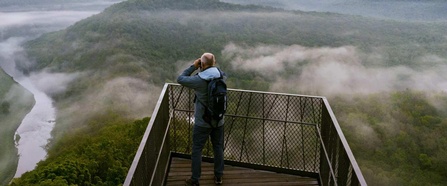 Mann blickt von einer Aussichtsplattform mit einem Fernglas über eine weitläufige, von Nebel durchzogene Wald- und Flusslandschaft.