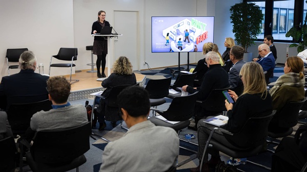 Eine Person steht am Rednerpult und präsentiert vor einem sitzenden Publikum; daneben zeigt ein großer Bildschirm eine RTR-Präsentationsfolie.