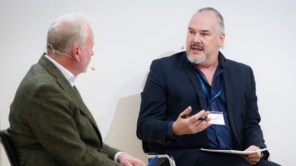 Zwei Personen sitzen in einem Gespräch auf dem Podium und diskutieren miteinander; beide tragen Headset-Mikrofone.
