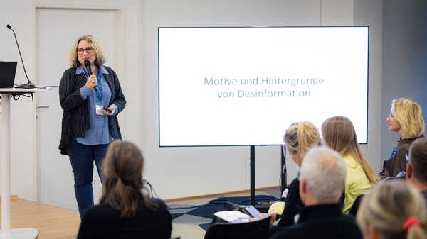 Eine Person steht auf dem Podium mit Mikrofon und spricht vor einem Publikum; auf dem Bildschirm ist der Titel „Motive und Hintergründe von Desinformation“ zu sehen.