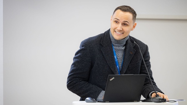 Eine Person steht am Rednerpult, lächelt und spricht in ein Mikrofon; vor sich ein Laptop und Unterlagen.