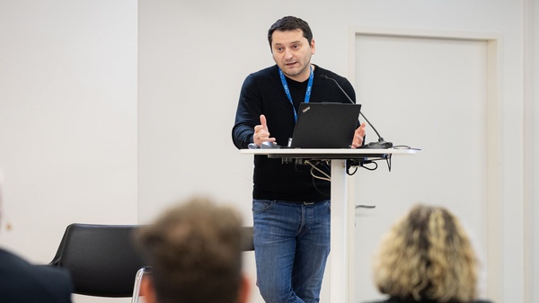 Eine Person steht am Rednerpult, spricht gestikulierend und blickt ins Publikum; vor sich ein Laptop und Mikrofone.