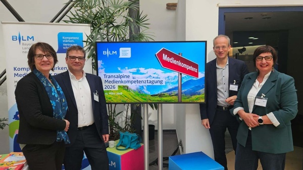Vier Personen posieren neben einem Bildschirm mit der Aufschrift „Transalpine Medienkompetenztagung 2026“ in einem Veranstaltungsbereich der BLM.