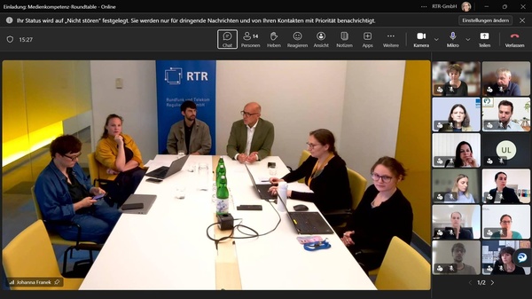 Mehrere Vertreter:innen der RTR-GmbH sitzen an einem Besprechungstisch und begrüßen die Teilnehmenden des Medienkompetenz-Roundtables. Die Veranstaltung findet hybrid statt, weitere Teilnehmende sind rechts in der Videokonferenz eingeblendet.