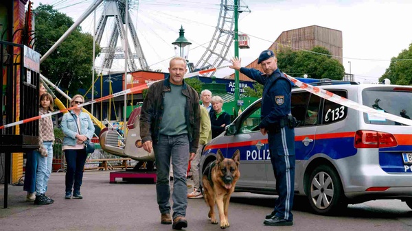 Mann geht mit einem Schäferhund durch einen abgesperrten Bereich im Wiener Prater; daneben steht ein Polizist vor einem Einsatzfahrzeug.