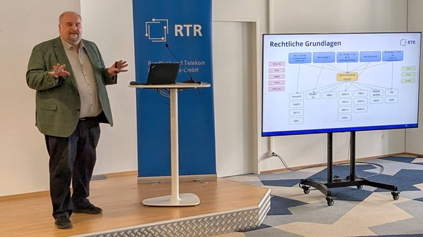 Magister Michael Ogris erläutert in einem Vortrag Aufgaben und Strukturen von KommAustria und RTR. Powerpoint-Folien werden auf einem großen Monitor angezeigt.
