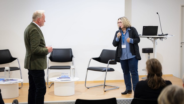 Zwei Personen stehen einander auf dem Podium gegenüber; eine Person spricht mit Mikrofon, die andere hört zu.