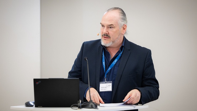 Eine Person spricht am Rednerpult und blickt leicht zur Seite, vor sich Unterlagen und ein Laptop.