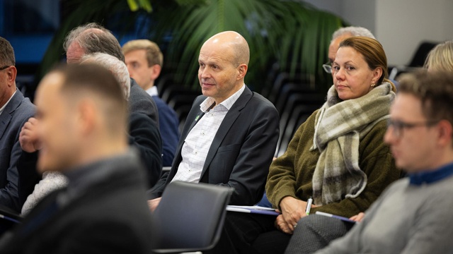 Mehrere Personen sitzen im Publikum und hören aufmerksam zu; manche machen sich Notizen.