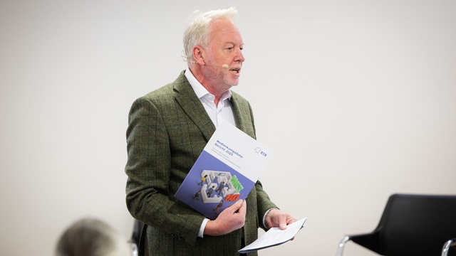 Eine Person hält den Medienkompetenzbericht 2025 in der Hand und spricht mit Headset-Mikrofon vor dem Publikum.