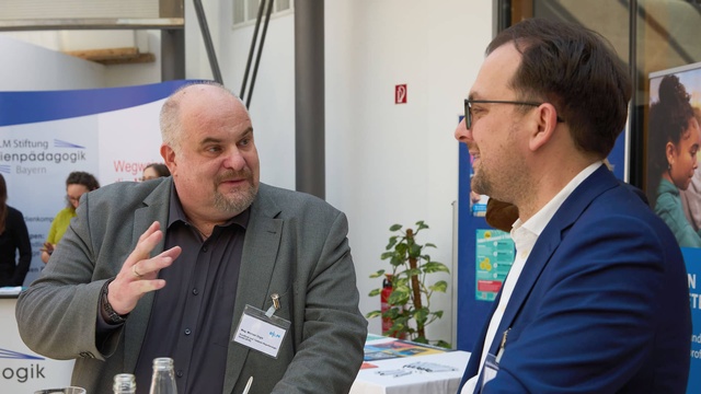 Zwei Männer unterhalten sich an einem Tisch im Foyer der Veranstaltung. Im Hintergrund sind Informationsstände und weitere Gäste zu sehen.