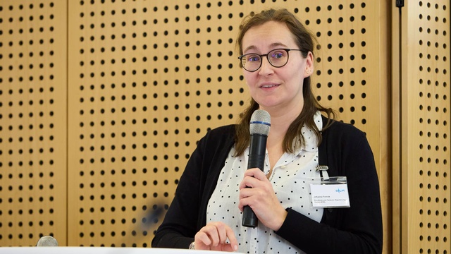 Eine Frau spricht mit Mikrofon bei einer Präsentation vor einer Holzpaneelwand.
