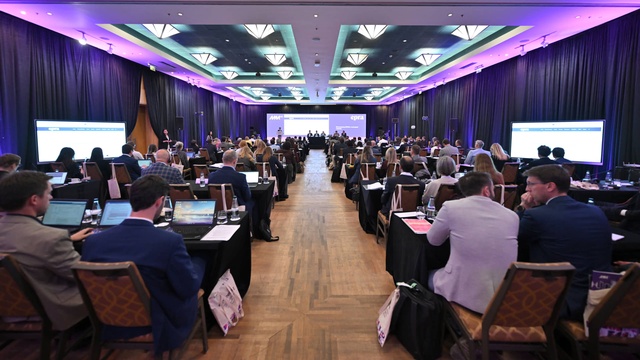 Weitwinkelaufnahme des Konferenzsaals mit zahlreichen Teilnehmenden an Tischen. Im Vordergrund ist die Bühne mit mehreren Vortragenden und einer großen Projektionsfläche zu sehen.