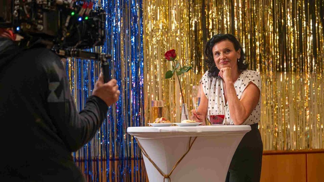 Frau sitzt mit einem Sektglas an einem festlich gedeckten Stehtisch vor einer goldenen Glitzerwand, während sie gefilmt wird.