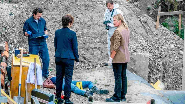 Ermittler:innen und Spurensicherung untersuchen einen Tatort auf einer Baustelle, an dem eine Person am Boden liegt.