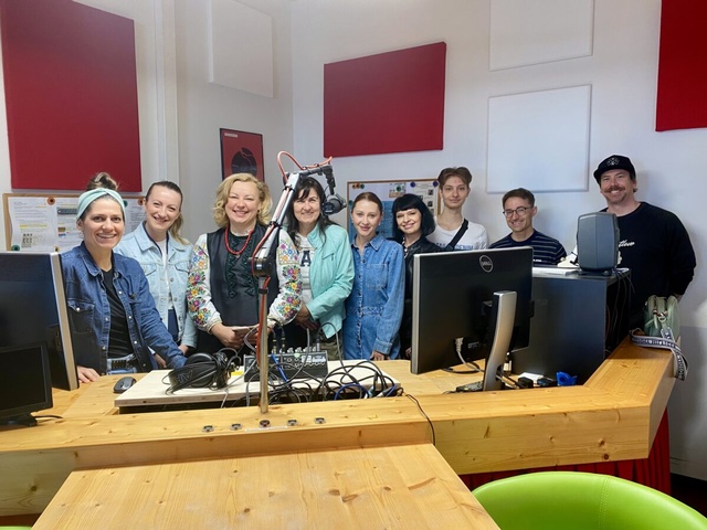 Mehrere Erwachsene stehen hinter einem Tisch mit Mischpult, Monitoren und Mikrofon in einem Radiostudio und blicken in die Kamera.