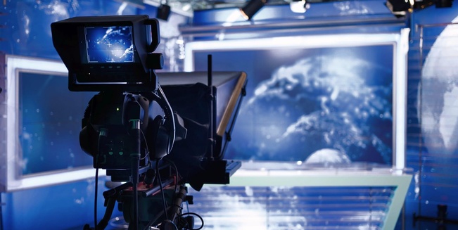 Themenbild Rundfunkförderungen. Im Vordergrund die Rückansicht einer Kamera im Fernsehstudio. Am Vorschaumonitor der Kamera erkennt man ein Moderationspult, das im Hintergrund des Fernsehstudios in der Unschärfe zu sehen ist.