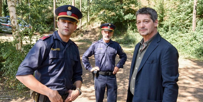 Links Schauspieler  Michael Steinocher als Polizist Martin und rechts Schauspieler Fritz Karl als Kommissar Paul Werner rechts, dazwischen im Hintergrund ein weiterer Polizist am Tatort im Wald.