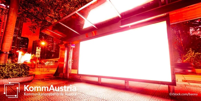 Das Themenbild für Veröffentlichungen und Ausschreibungen der KommAustria zeigt eine moderne Bushaltestelle umgeben von Bäumen und Häusern bei Nacht. In dem überdachten Unterstand leuchtet hellweiß eine leere Werbefläche.