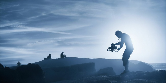 Ein Mann steht in einer Lavalandschaft und hält eine mittelgroße Filmkamera in Händen, die er auf zwei Personen am Horizont richtet. Das Bild ist insgesamt blau eingefärbt. 
