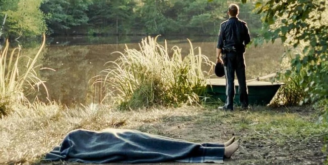Polizist Martin, gespielt von Michael Steinocher, steht mit dem Rücken zum Betrachter und blickt allein über den Teich im Wald. Im Vordergrund liegt am weiteren Ufer die abgedeckte Leiche.