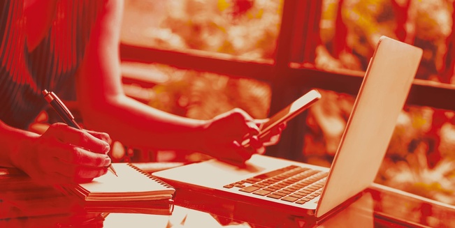 Frauenhände, eine Hand mit Stift, die andere mit Handy, davor ein Laptop