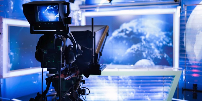 Themenbild Rundfunkförderungen. Im Vordergrund die Rückansicht einer Kamera im Fernsehstudio. Am Vorschaumonitor der Kamera erkennt man ein Moderationspult, das im Hintergrund des des Fernsehstudios in der Unschärfe zu sehen ist.