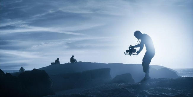 Ein Mann steht in einer Lavalandschaft und hält eine mittelgroße Filmkamera in Händen, die er auf zwei Personen am Horizont richtet. Das Bild ist insgesamt blau eingefärbt. 