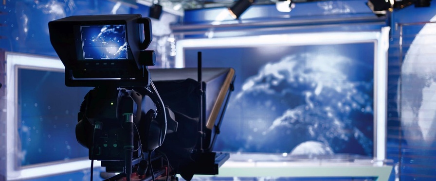 Themenbild Rundfunkförderungen. Im Vordergrund die Rückansicht einer Kamera im Fernsehstudio. Am Vorschaumonitor der Kamera erkennt man ein Moderationspult, das im Hintergrund des Fernsehstudios in der Unschärfe zu sehen ist.