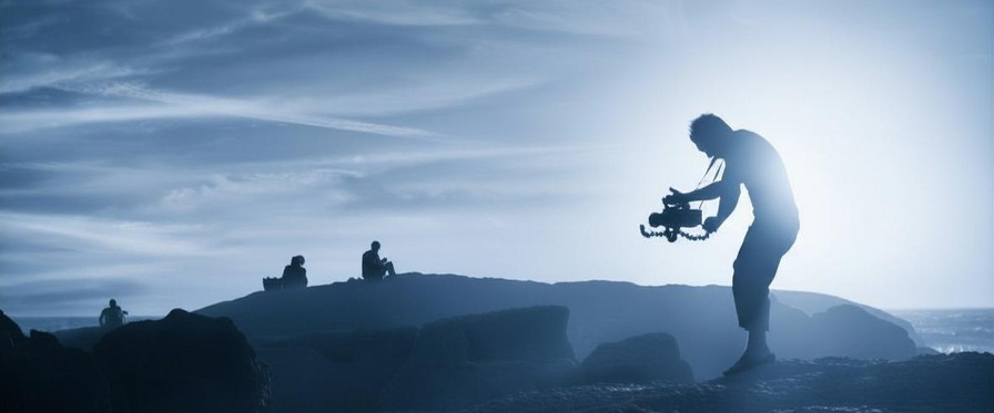 Ein Mann steht in einer Lavalandschaft und hält eine mittelgroße Filmkamera in Händen, die er auf zwei Personen am Horizont richtet. Das Bild ist insgesamt blau eingefärbt. 
