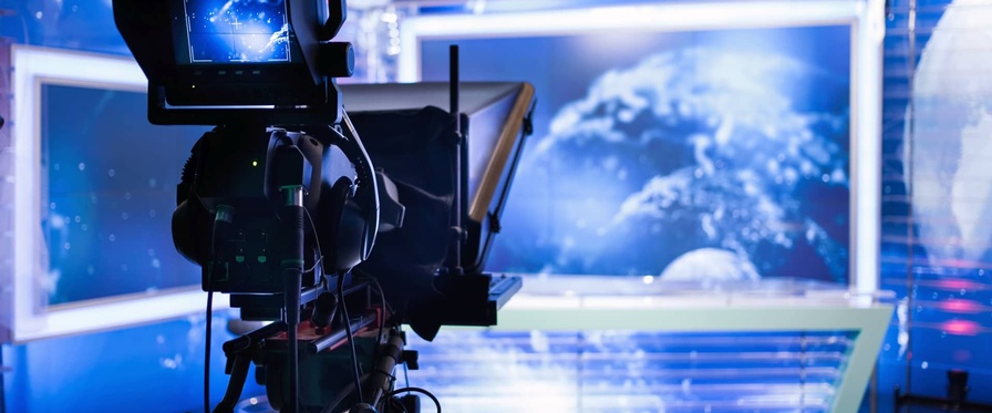 Themenbild Rundfunkförderungen. Im Vordergrund die Rückansicht einer Kamera im Fernsehstudio. Am Vorschaumonitor der Kamera erkennt man ein Moderationspult, das im Hintergrund des des Fernsehstudios in der Unschärfe zu sehen ist.