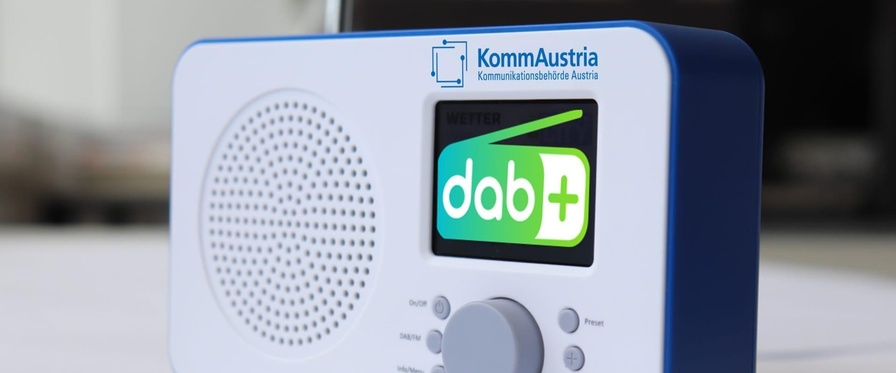 Kleines, tragbares Radiogerät in Weiß mit blauem Rahmen und Aufdruck KommAustria sowie dem Logo von DAB plus am Display
