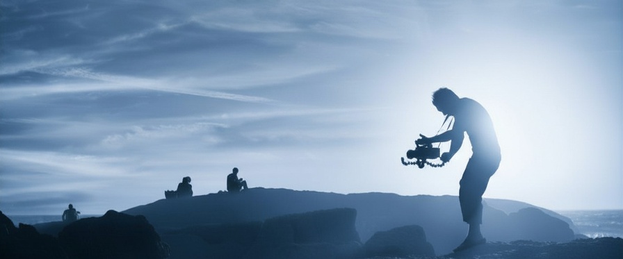 Ein Mann steht in einer Lavalandschaft und hält eine mittelgroße Filmkamera in Händen, die er auf zwei Personen am Horizont richtet. Das Bild ist insgesamt blau eingefärbt. 