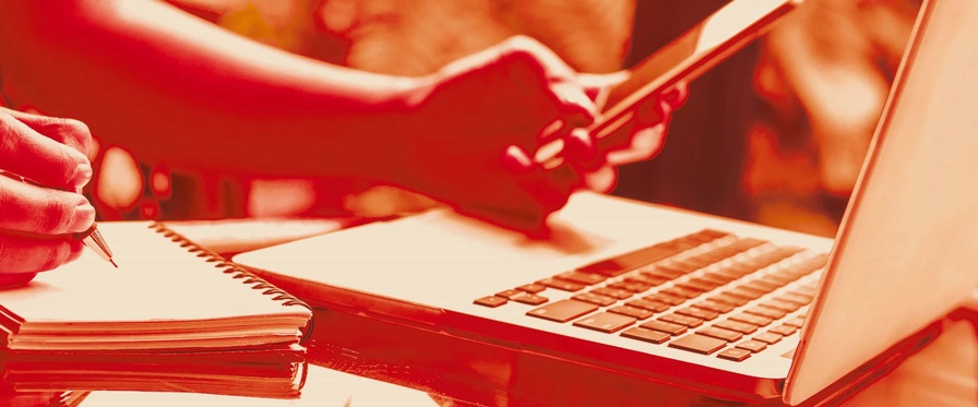 Frauenhände, eine Hand mit Stift, die andere mit Handy, davor ein Laptop